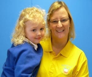 Helen Webster, a Talkfirst Franchisee, smiles in a yellow shirt while holding a young child in a blue sweater against a blue background.