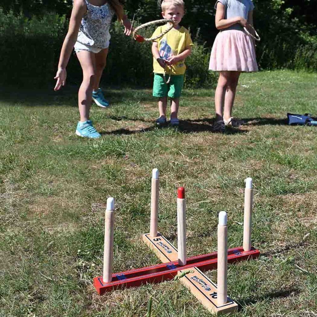 Three children play a ring toss game on a grassy lawn, aiming to throw rings onto wooden pegs labeled with point values—each hopeful toss like taking the first steps toward a future career.