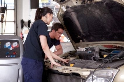 Two mechanics balancing work and family life while working on a car in a garage.