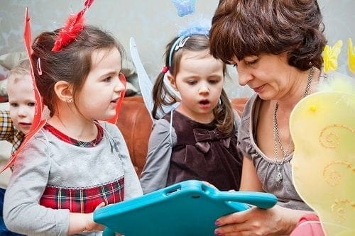 A woman, balancing her career and work-life responsibilities, is holding a tablet while a group of children look at it.