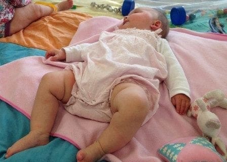 A baby is laying on a pink blanket in a room full of toys while their mum enjoys a moment of work-life balance.