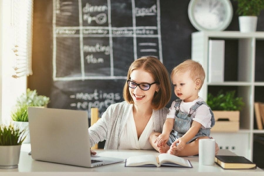 A mum is working on a laptop with a baby in front of it.