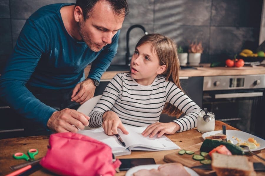 A man and a girl working on homework in the kitchen while the mum, a woman returner, is working.
