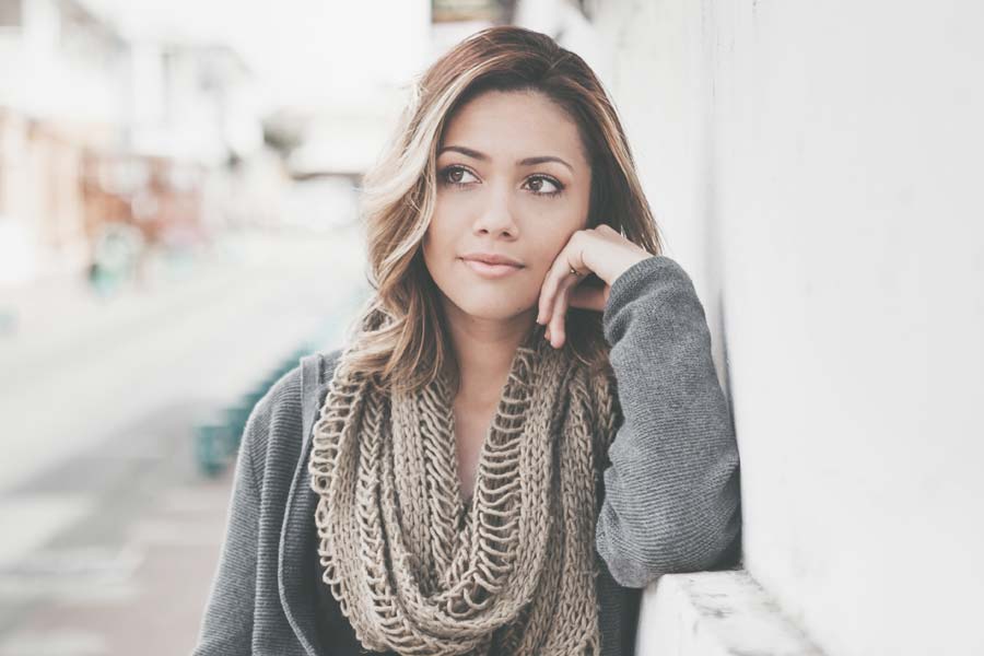 A woman with wavy hair and a knitted scarf leans against a white wall, looking thoughtfully into the distance—perhaps reflecting on her career or balancing the demands of jobs and mums.