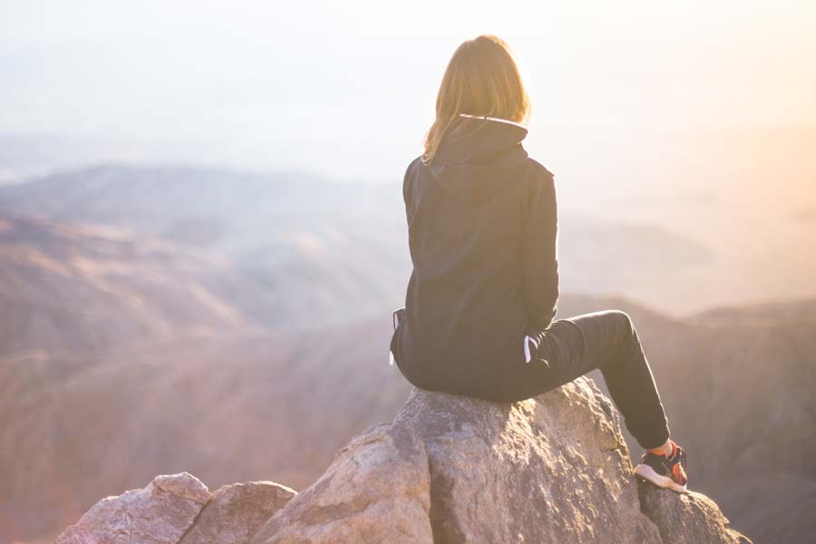 A person wearing a black hoodie and sneakers, perhaps reflecting on work or career, sits on a rock facing a hazy mountain landscape under soft sunlight.