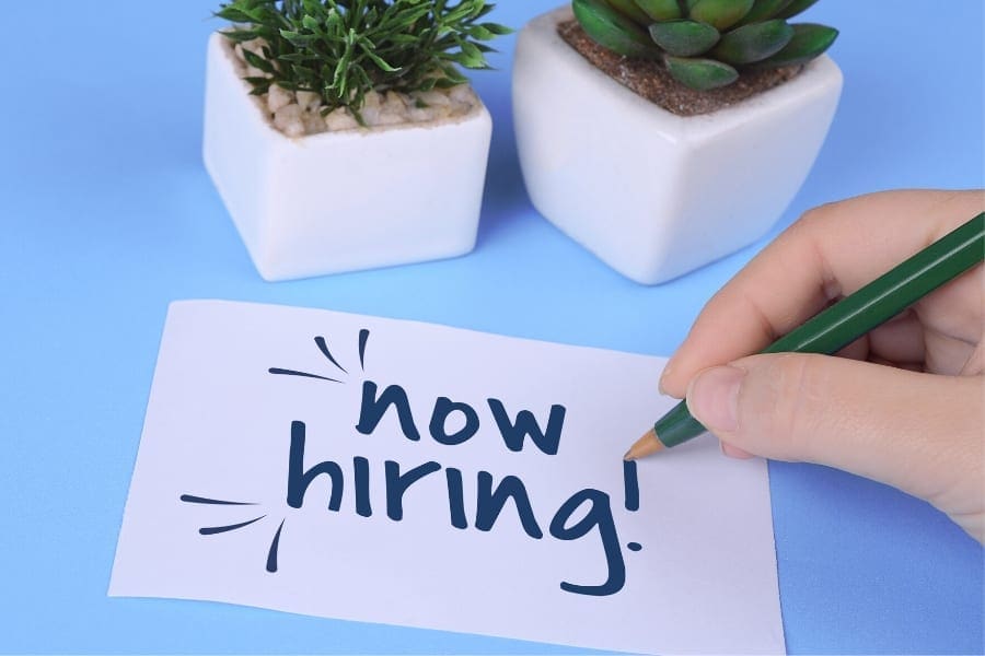 An individual writing the word "now hiring" on a piece of paper, specifically targeting women returners or moms seeking work.