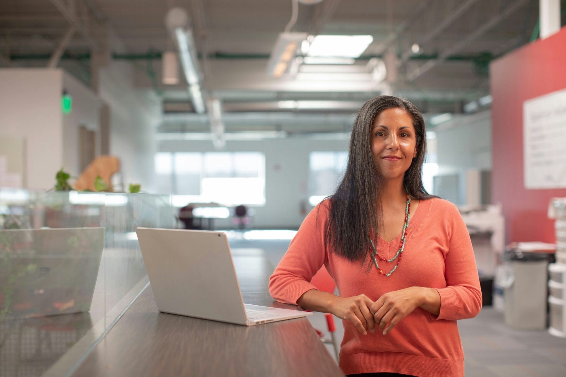 A woman with long dark hair, wearing an orange top, sits at a desk with a laptop in a modern office space, smiling at the camera—reflecting UK business efforts to boost employee retention after the Great Resignation.