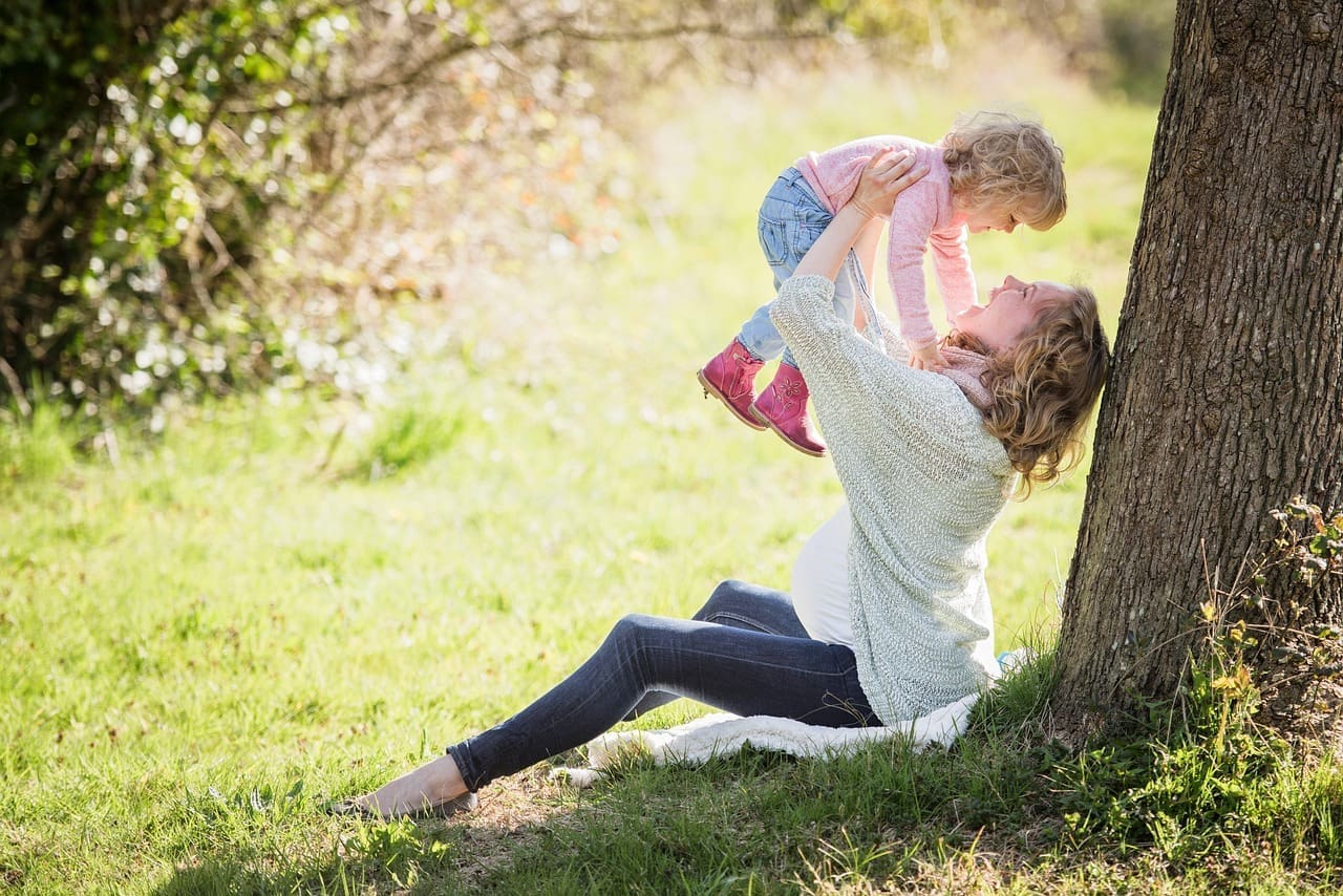 A woman sits against a tree in a grassy area, lifting a young child above her. Both appear to be smiling and enjoying time outdoors, embracing the joys of raising kids and finding work-life balance.