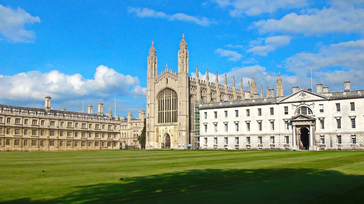 A large Gothic-style chapel and historic college buildings stand behind a wide green lawn under a blue sky with scattered clouds, evoking the timeless allure of college admission to a dream university.