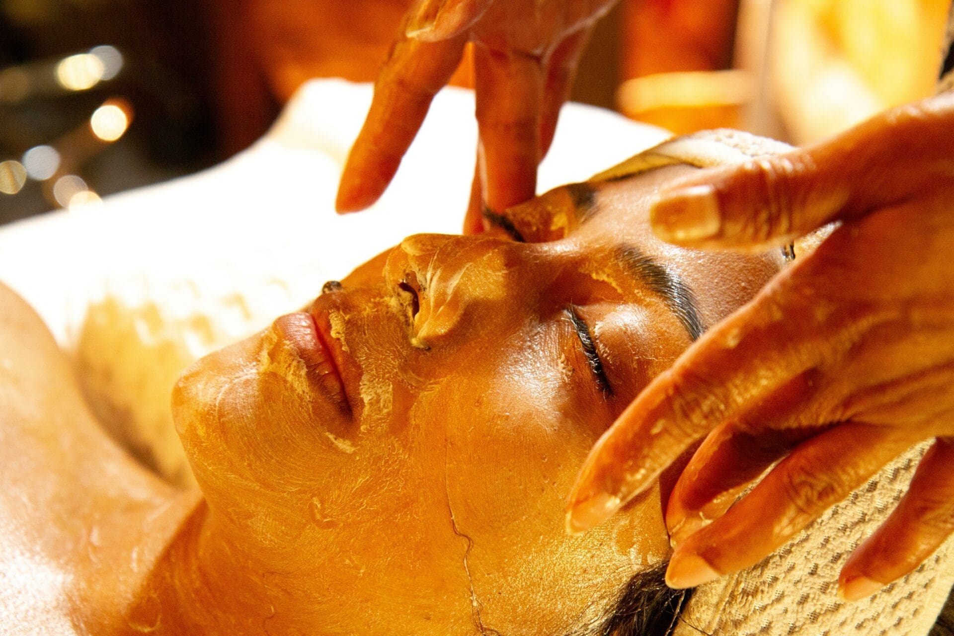 A person with eyes closed receives a facial treatment featuring a yellowish mask, while a professional—likely trained through skincare diploma programs—gently massages their forehead for relaxation and revitalization.