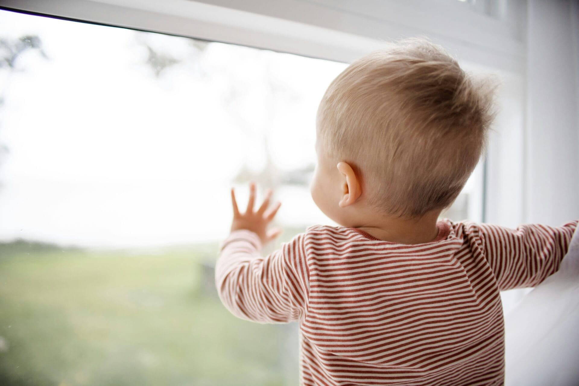 A young child with short blond hair wearing a striped shirt looks outside and touches a window with one hand, imagining adventures beyond—a reminder of why families seek ways to beat childcare costs.