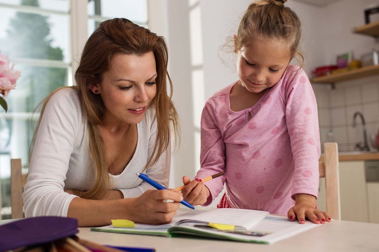 An adult woman provides homework help to a young girl at a table in a bright kitchen, both focused on an open notebook.