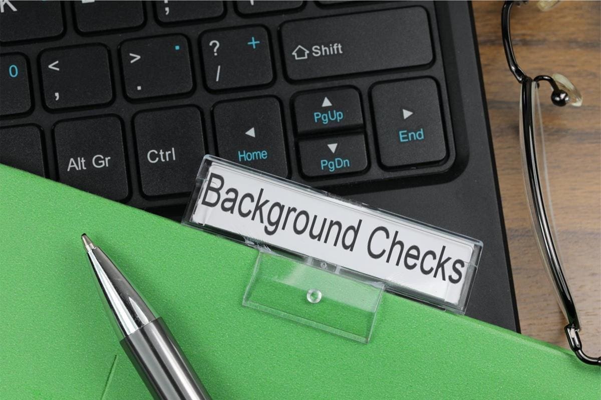A nameplate labeled "Background Checks" sits on a green folder next to a pen, glasses, and a computer keyboard—essential tools for hiring checks when you’re ready to hire someone.
