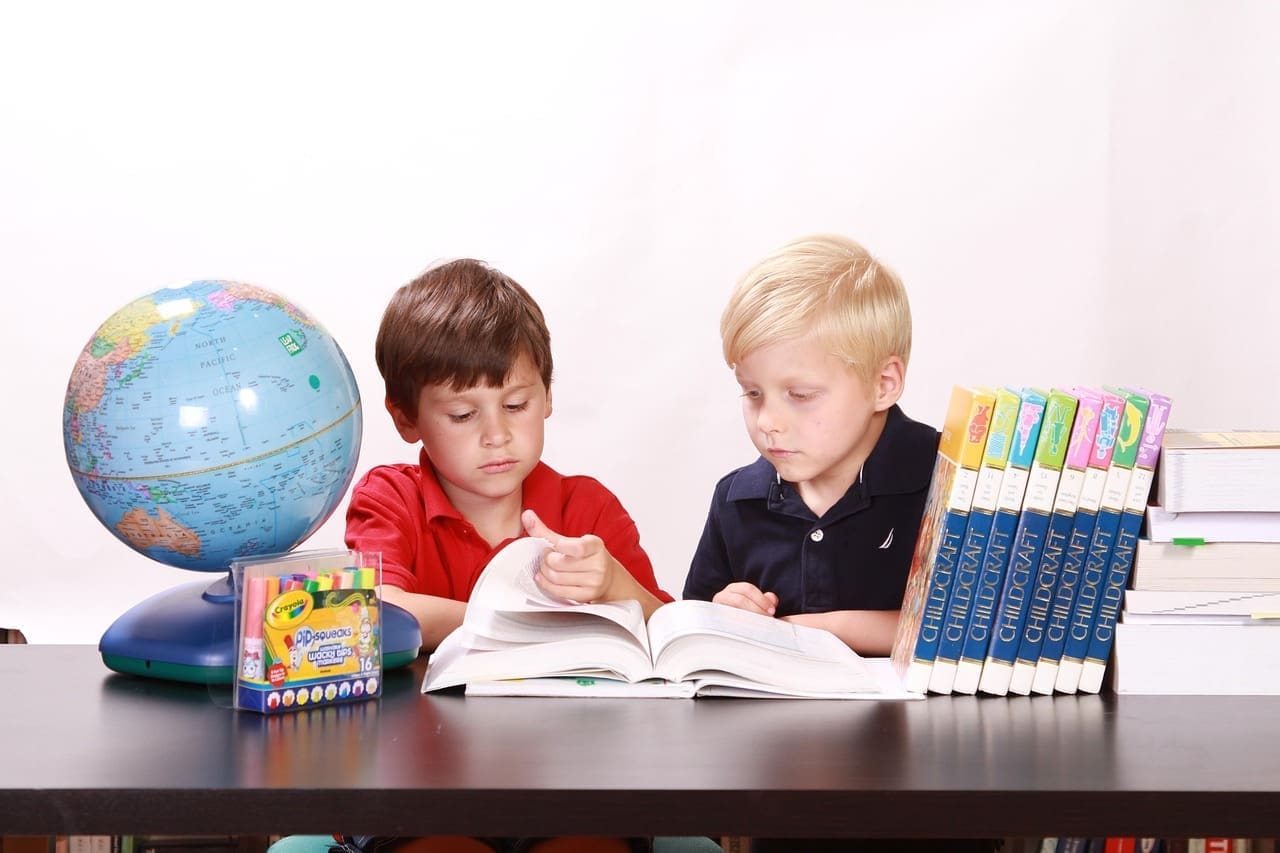 Two young boys sit at a table studying a book, with a globe, crayons, and a stack of dictionaries beside them, eager to make the most of education.