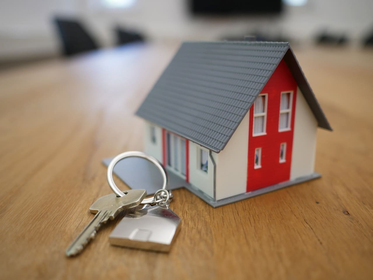 A small model house with a red front and gray roof sits on a wooden table next to silver house keys, symbolizing the dream of home buying for any busy professional.