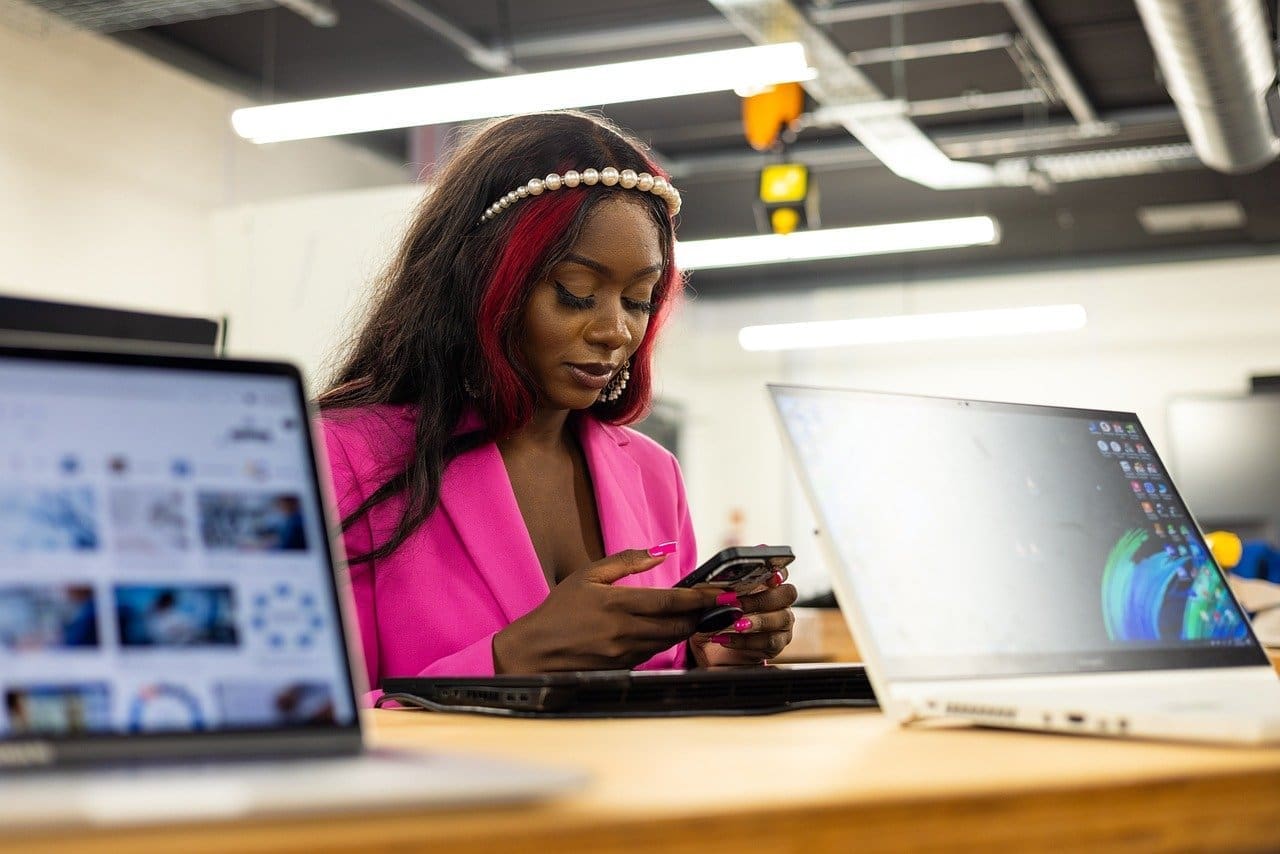 A woman in a bright pink blazer uses her phone at a desk with two open laptops in a modern office environment.