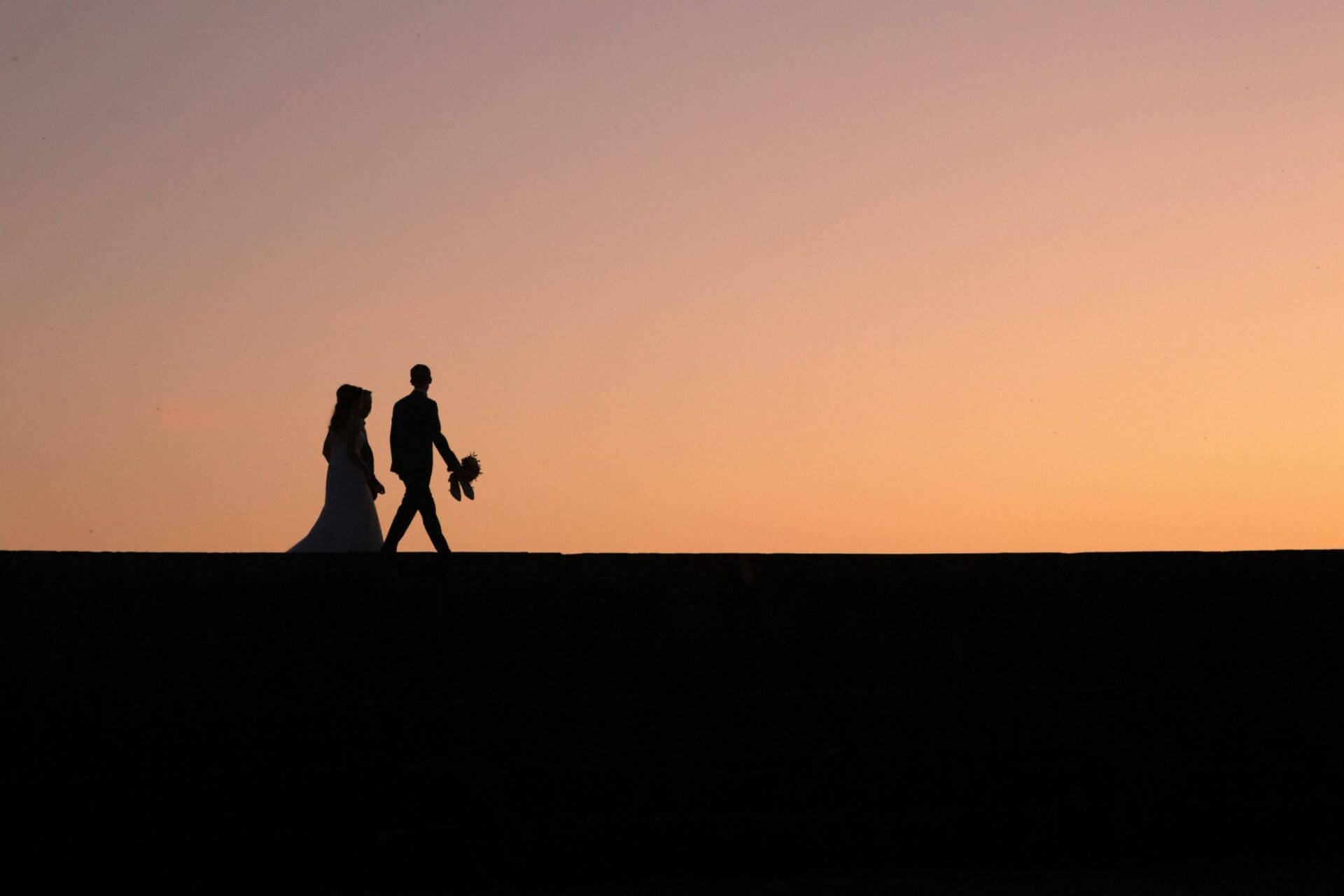 Silhouette of a bride and groom walking on a flat surface against an orange sunset sky, symbolizing marriage stability and the journey couples take together.