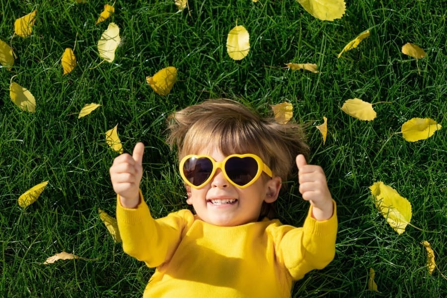 A child in a yellow shirt and heart-shaped sunglasses lies on the grass in an outdoor play area, surrounded by yellow leaves, smiling and giving two thumbs up.