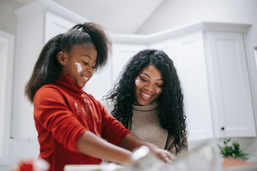 Two people in a kitchen, one wearing a red shirt and the other a beige sweater, with flour on their faces and hands. Smiling together, they appear to be baking or cooking. It's a perfect scene when cooking with kids, highlighting the fun and benefits for children in the kitchen.