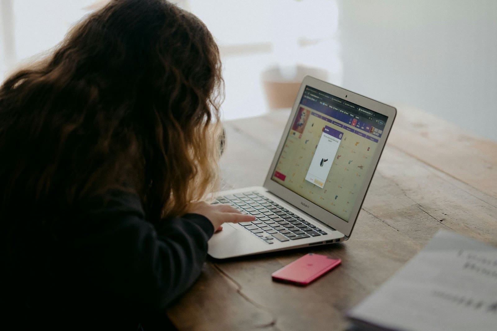 A person with long hair uses a laptop at a wooden table, with a pink phone and papers nearby. The laptop screen displays a game or quiz about safe internet use, highlighting important internet tips for kids.