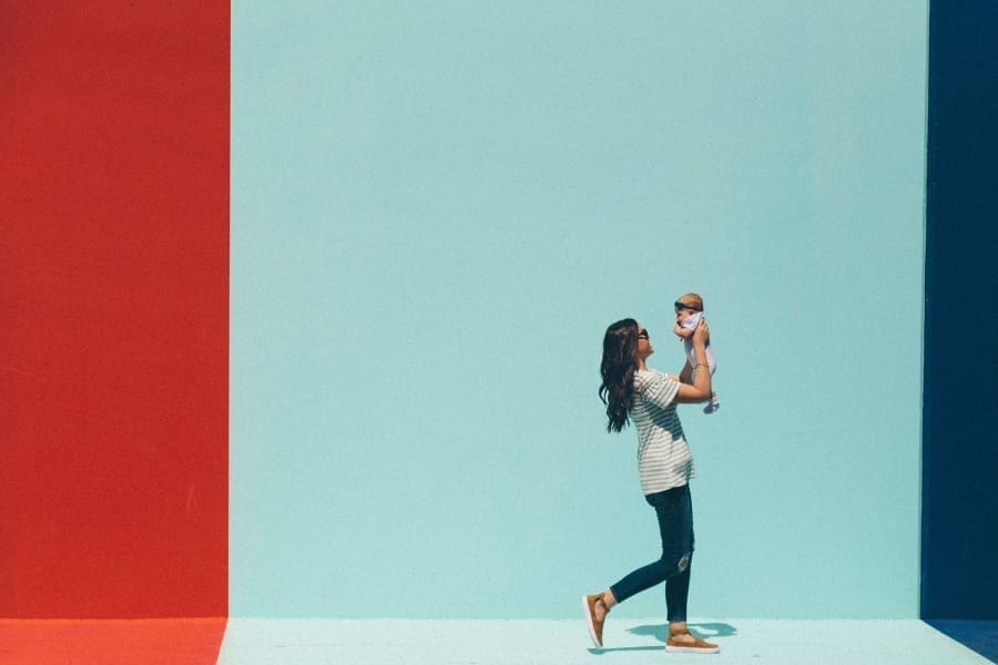 A busy mom holds a baby up against a colorful backdrop with red, light blue, and navy vertical stripes, embodying everyday moments filled with love and vibrant smiles.
