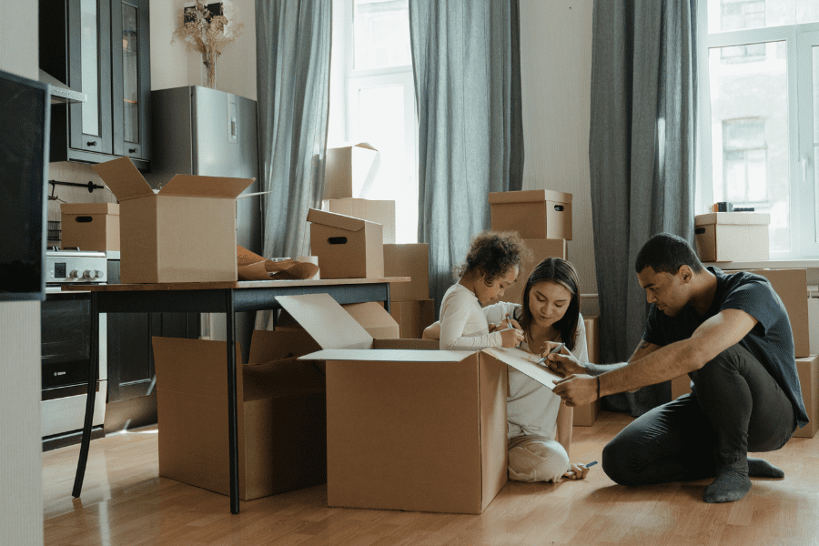 A family of three sits among cardboard boxes in a living room, sorting or packing items, suggesting a relocation adventure with all its logistics challenges.