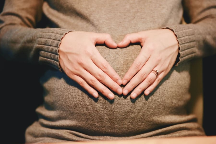 A person in a gray sweater forms a heart shape with their hands over their pregnant belly, embodying the essence of best maternal care.