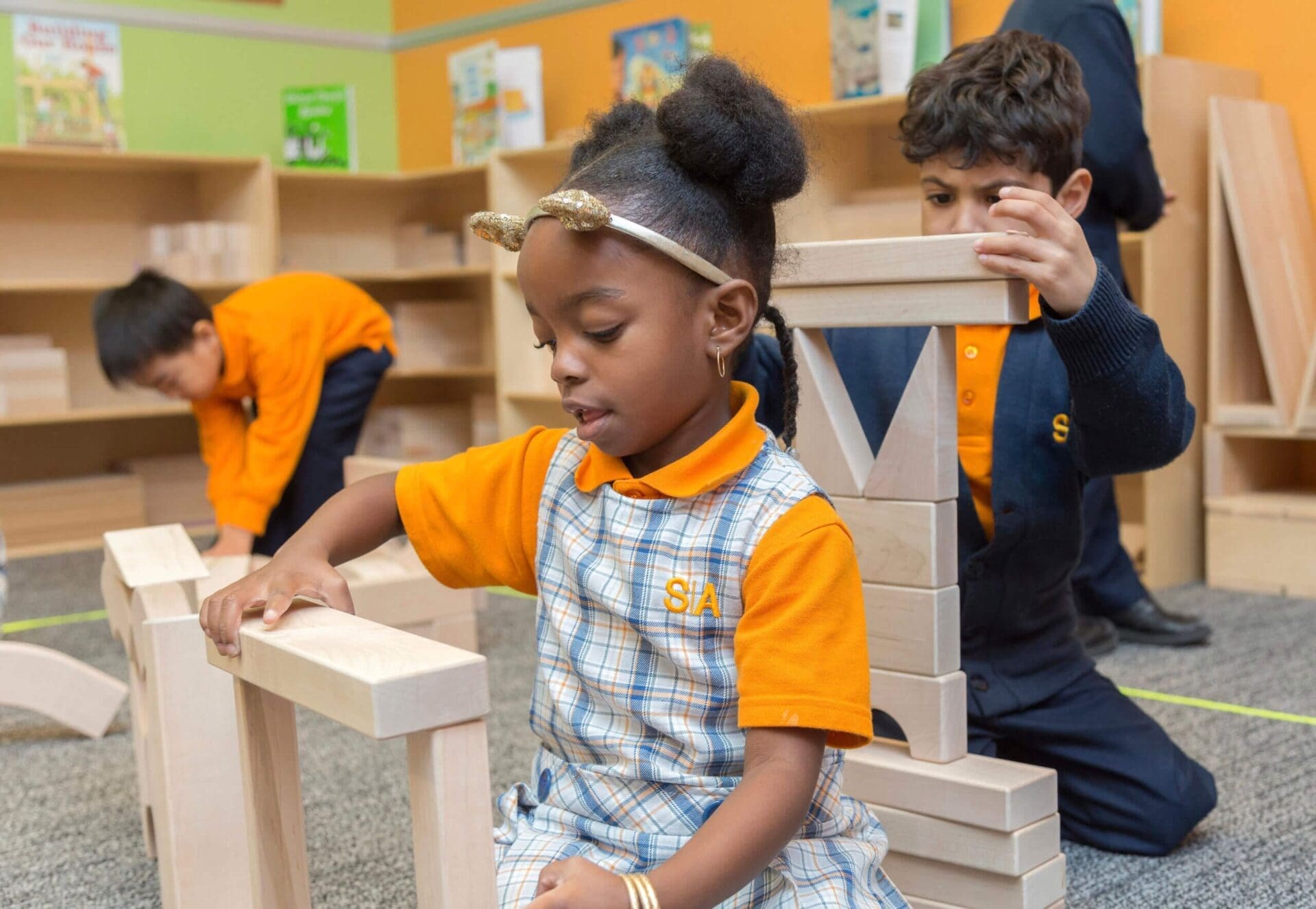 Three young children in uniforms build structures with wooden blocks in a classroom setting.