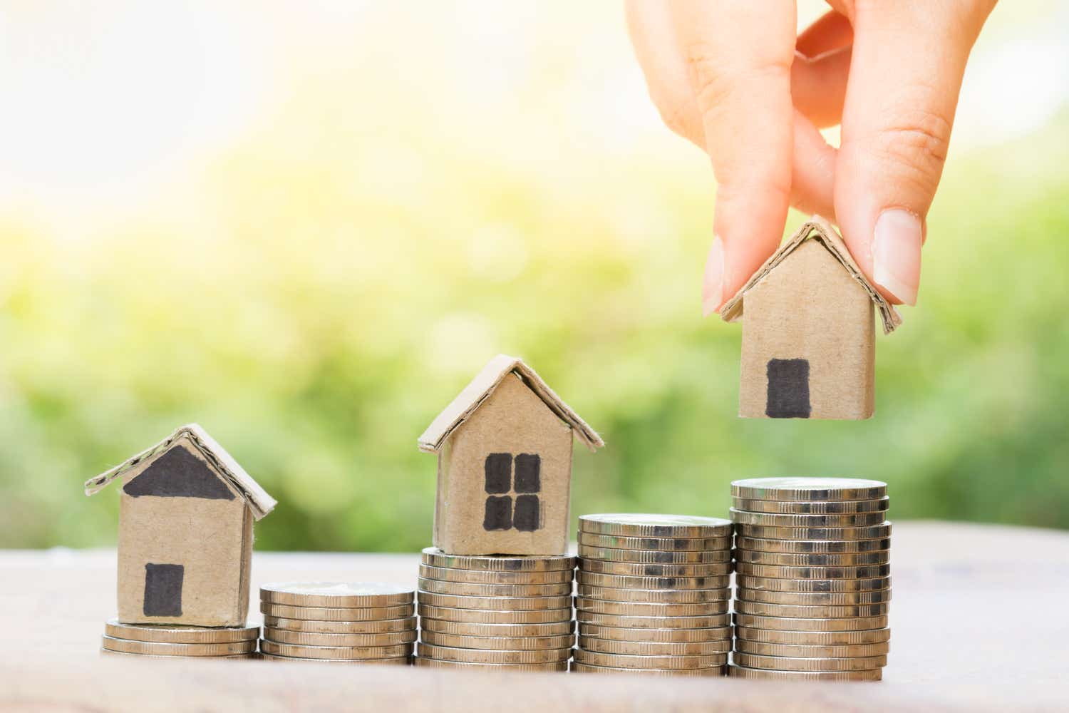 A hand places a small cardboard house on top of stacked coins, with two other small houses on stacks of coins beside it, symbolizing real estate investment or property value.