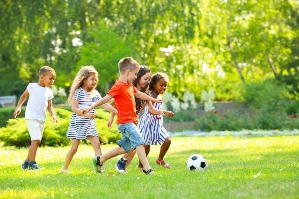 Five children play soccer on a grassy field in a park on a sunny day, with trees and plants in the background.