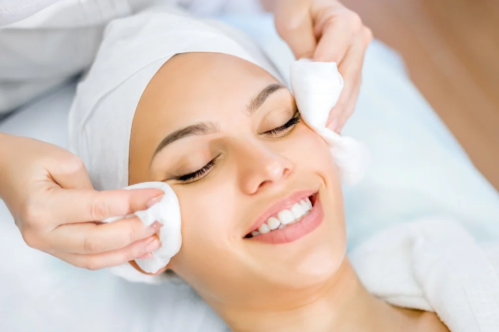 A person with a headband is lying down and smiling while receiving a facial treatment, as someone gently cleanses their face with cotton pads.
