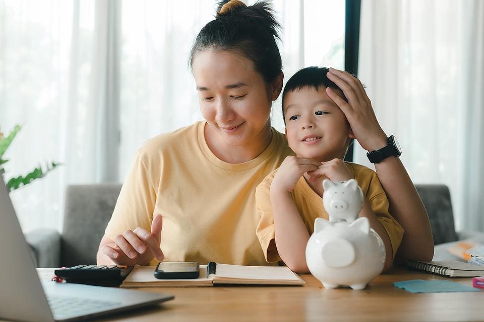 A woman and a young boy sit at a table with a notebook, calculator, and piggy bank, looking at a laptop screen together in a bright room.