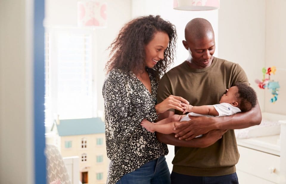 Two adults standing in a nursery, smiling and looking at a baby they are holding together.