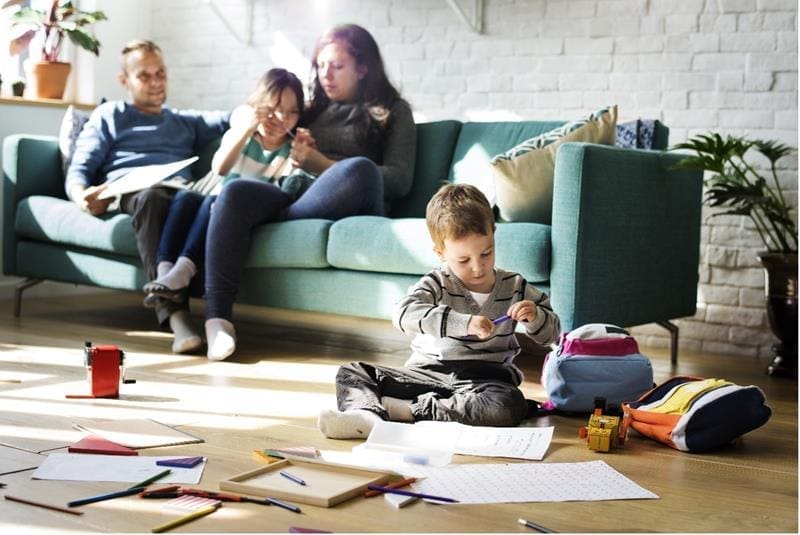 A young boy sits on the floor drawing amid scattered papers, while three people sit on a sofa in the background, engaged in various activities.