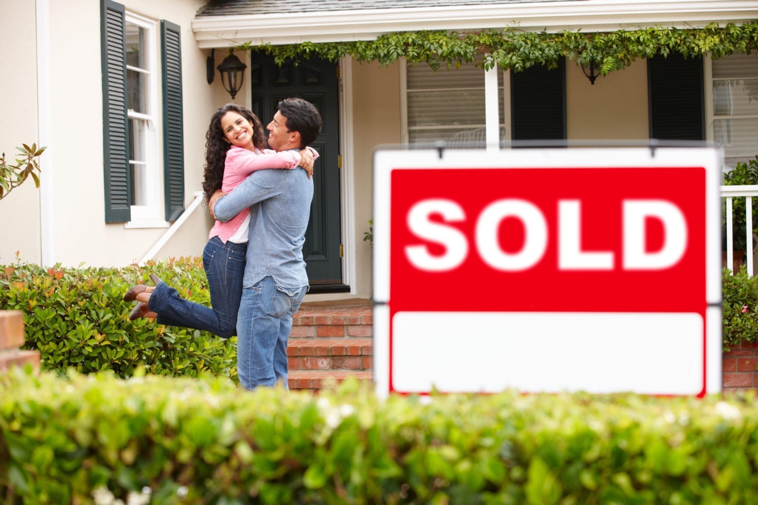 A couple embraces and smiles in front of a house with a "SOLD" sign in the yard. The home has white siding, green shutters, and a brick porch.