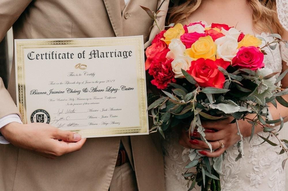 A couple in wedding attire holds a certificate of marriage and a bouquet of red, pink, yellow, and white roses.