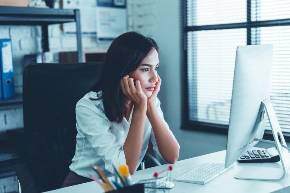 A woman sits at a desk, resting her face in her hands, and looks at a computer monitor in an office setting.