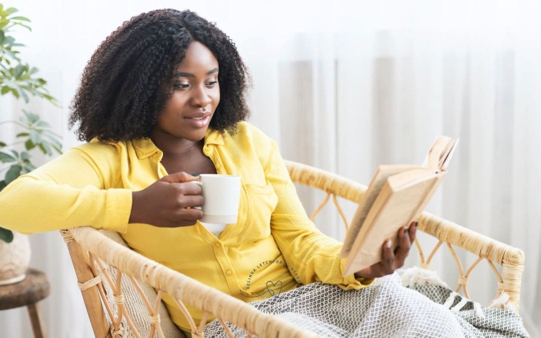 Woman sitting in a wicker chair, wearing a yellow shirt, holding a white mug and reading a book, with a blanket on her lap and a plant in the background.