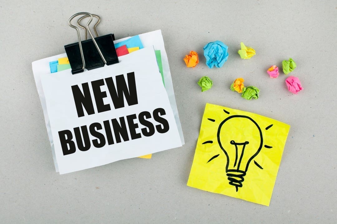 A stack of papers clipped together with "NEW BUSINESS" written on top, next to a yellow sticky note with a drawing of a light bulb and several crumpled paper balls.
