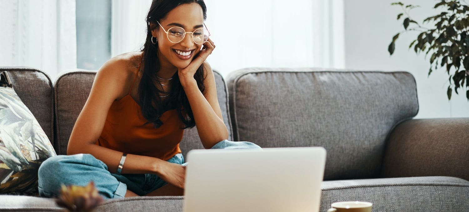 Woman sitting cross-legged on a couch, smiling while looking at a laptop. A coffee cup is on the table and a plant is in the background.