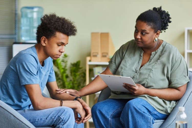 A therapist holding a clipboard offers support to a teenage boy, who looks sad and is sitting with his head down.