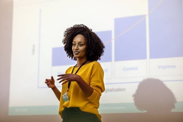 A woman in a yellow sweater is giving a presentation in front of a projected bar graph.