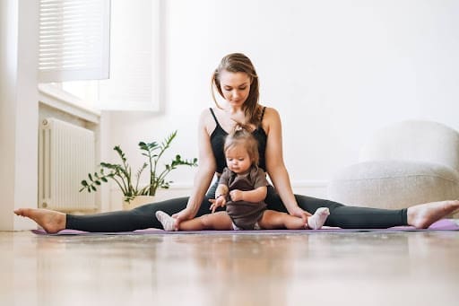 A working mom practices self-care as she sits on a yoga mat in a wide-legged stretch, her toddler seated in front of her. Both face forward, sharing a bright, peaceful moment together.