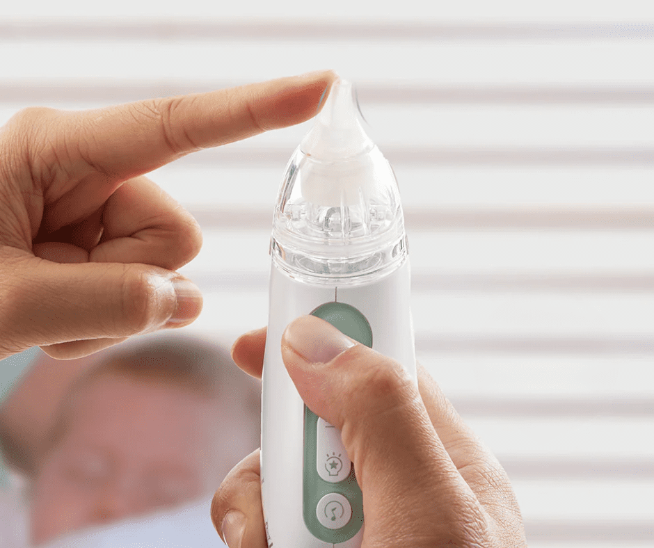 A person holds and presses the tip of a white and green electronic nasal aspirator, removing mucus as a blurred infant sits in the background—a helpful tool from any parent's guide to care.